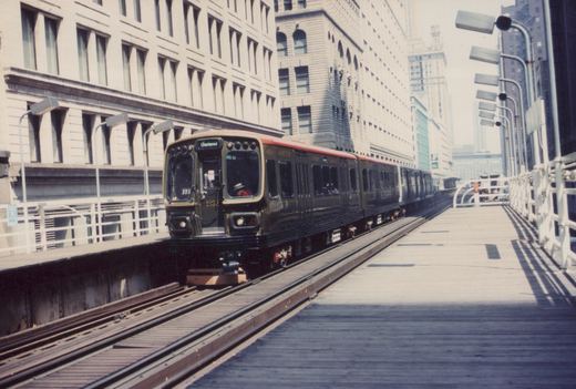 A dark brown and cream Chicago Transit Authority rapid transit train, number 1892, is stopped at a station on an elevated track in a city, with a conductor visible in the front window and tall buildings in the background.