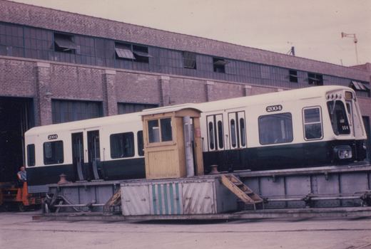 A white and green CTA rapid transit car, number 2004, sits on a flatcar in front of a brick building, with an orange forklift and a man nearby.