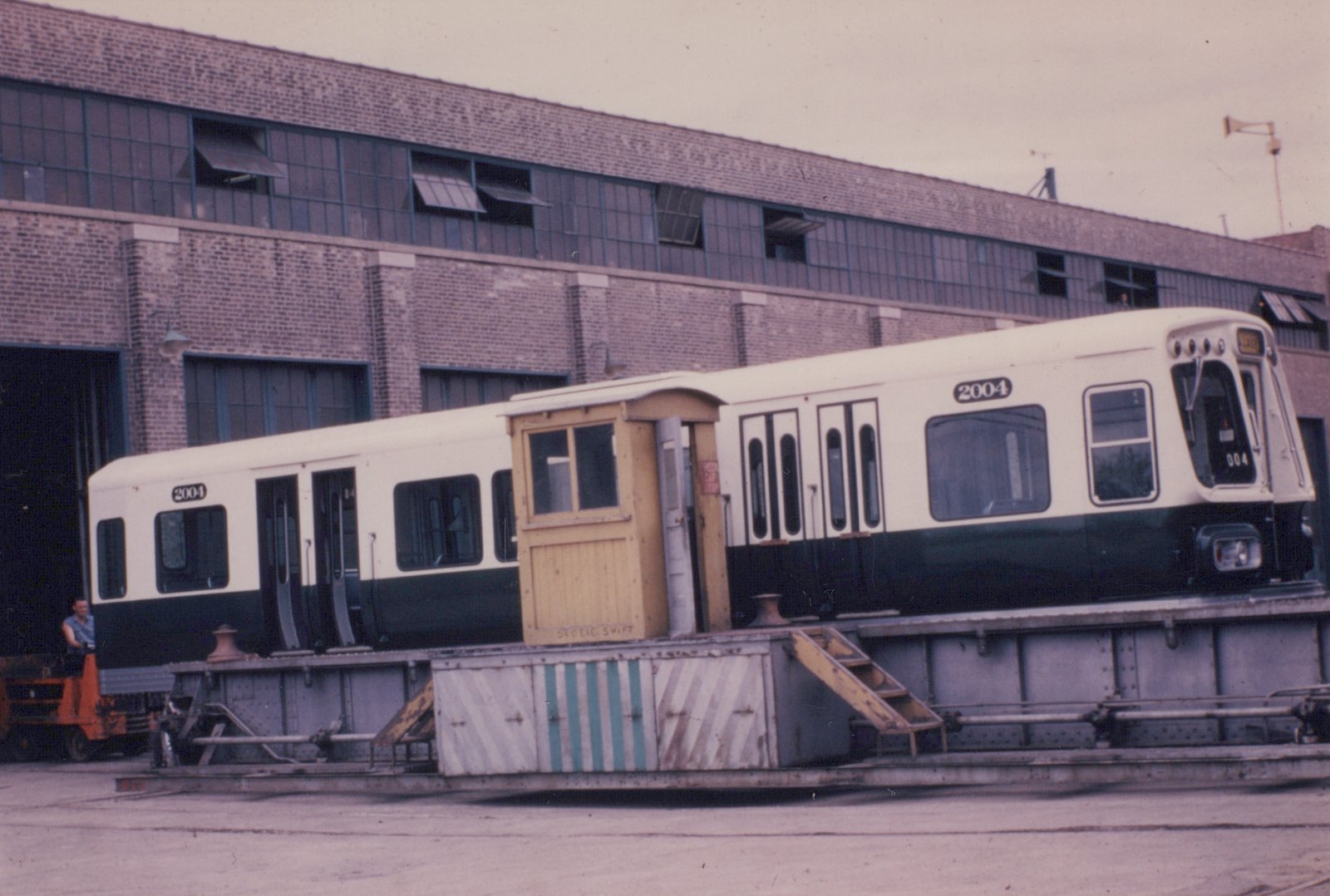 A white and green CTA rapid transit car, number 2004, sits on a flatcar in front of a brick building, with an orange forklift and a man nearby.