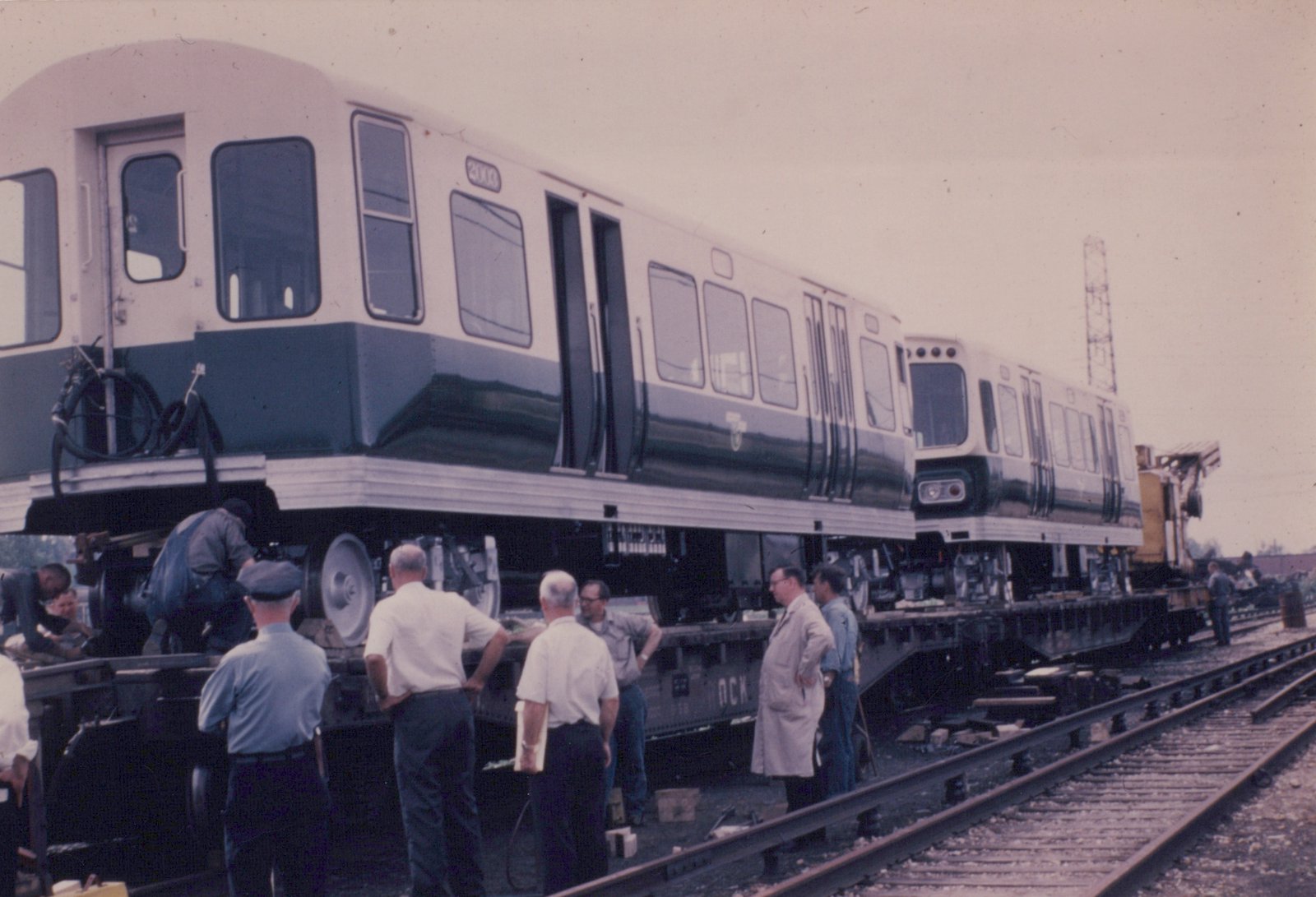 A white and dark-colored rapid transit car, numbered 2003, sits on a flatcar in a rail yard, surrounded by several men observing and working on it.