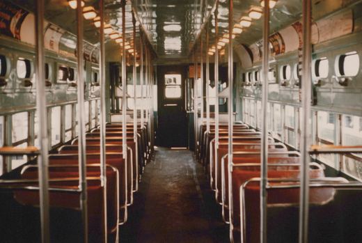 An empty vintage passenger car interior with rows of reddish-brown seats, chrome poles, and large windows, viewed from the aisle looking toward the far door.