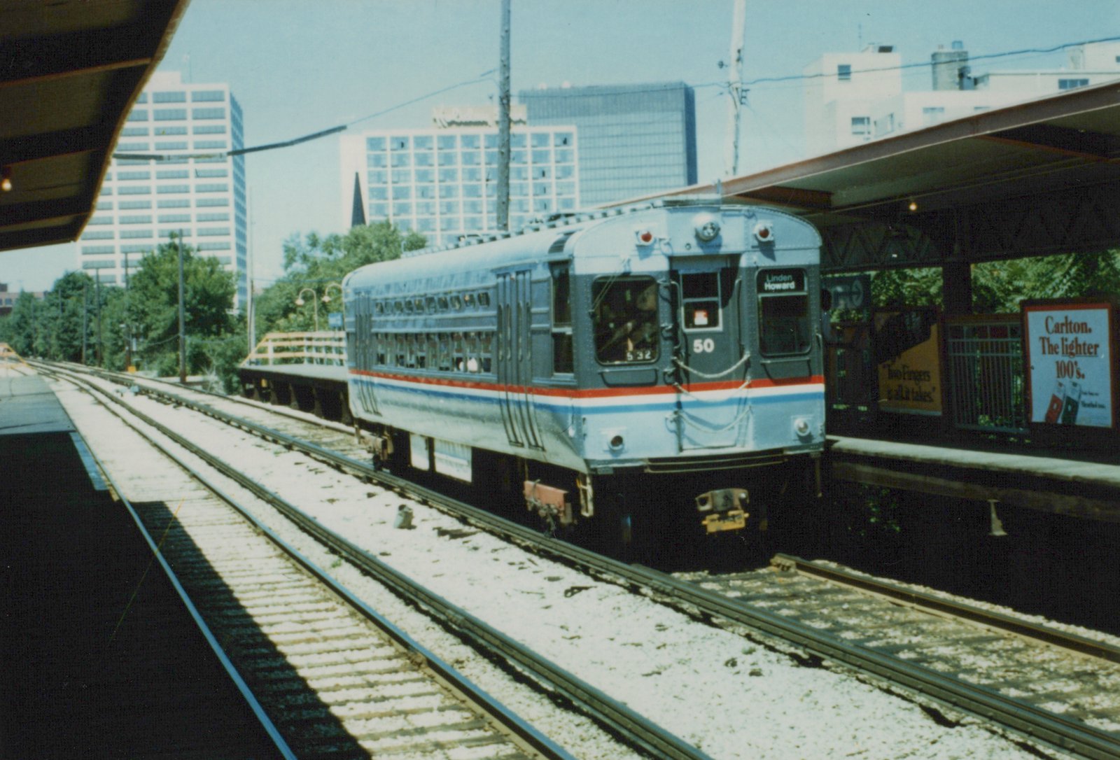 A silver and blue electric railcar with the number 50 on the front, stopped at a station platform under a clear sky, with city buildings in the background.