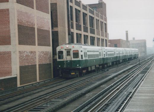 A green and cream CTA train, number 47, on an elevated track beside a large brick building under an overcast sky.