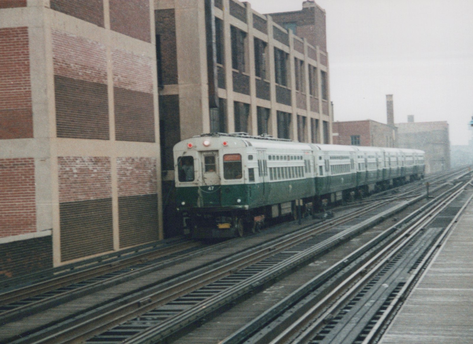 A green and cream CTA train, number 47, on an elevated track beside a large brick building under an overcast sky.