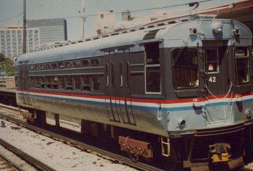 A single electric multiple-unit train car, number 42, with a red, white and blue stripe, sits on elevated tracks under a clear sky, with a 'Linden Howard' sign in its front window and a Holiday Inn in the background.