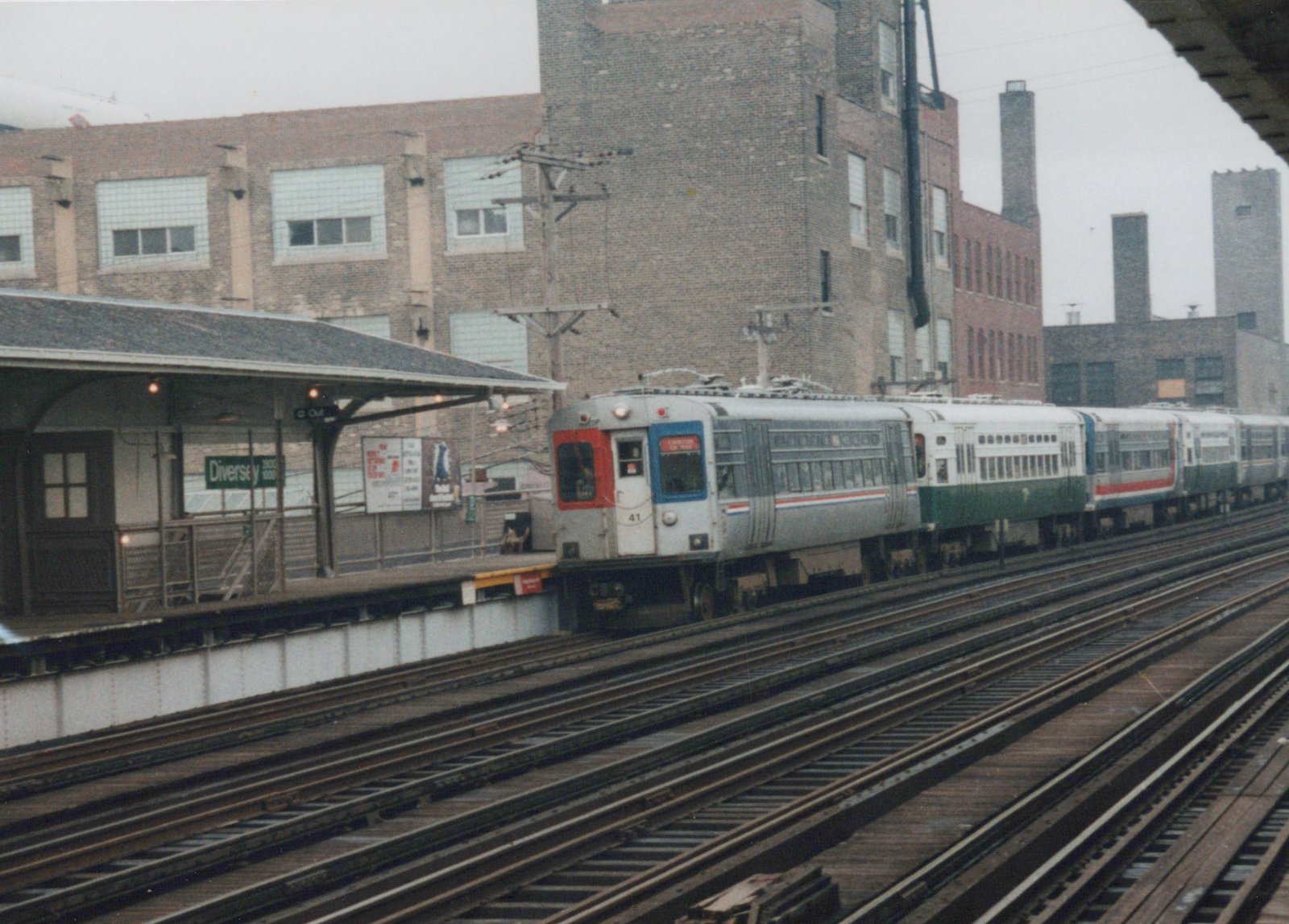 A silver, green, and red electric train with the number 41 is stopped at the Diversey elevated station, with industrial buildings in the background.