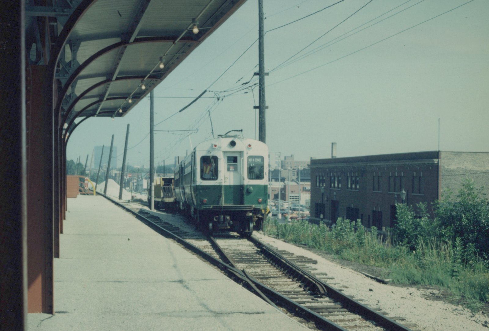 A green and white electric multiple-unit train, number 406, marked 'NOT IN SERVICE', is stopped on an elevated track beside a station platform, with overhead power lines and a brick building in the background.