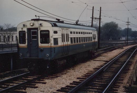 A blue and white electric multiple-unit train car on elevated tracks under a gray sky, with the destination sign 'HOWARD' visible on the front.