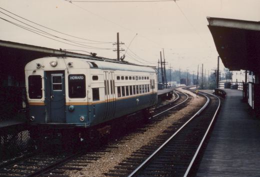 A two-toned CTA train car with the destination 'HOWARD' displayed, stopped at a wooden platform on an elevated track under an overcast sky.