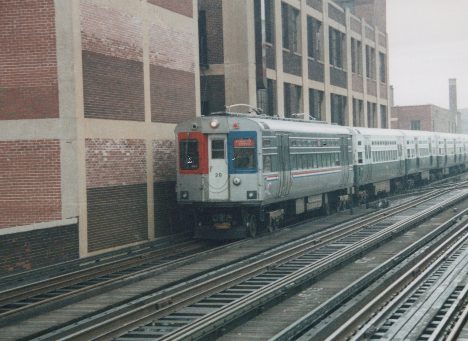 A gray and red CTA rapid transit train on elevated tracks next to a brick building, with the number 28 and destination 'Evanston Express' visible.