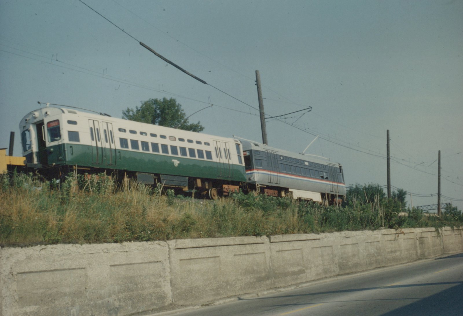 Two electric railcars, one green and white and one silver with red and blue stripes, are on a track beside a highway with a concrete barrier.