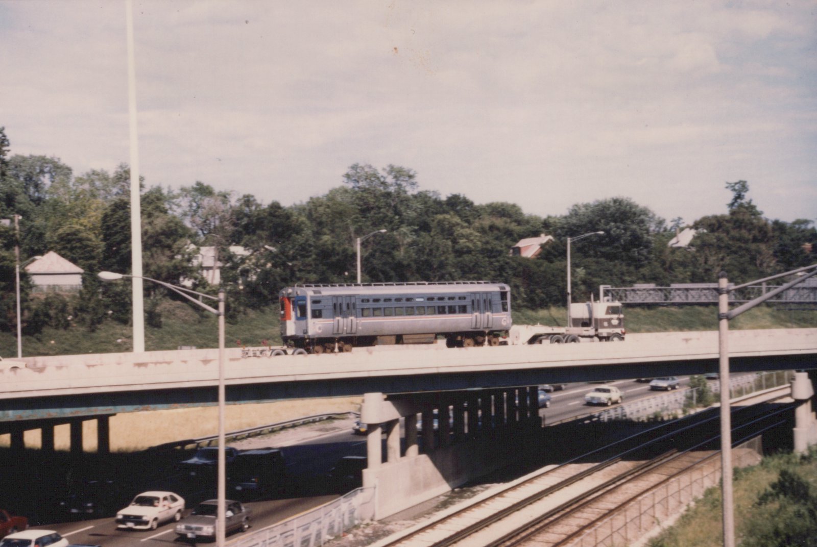 A silver CTA train car on an elevated track above a busy highway, with trees and houses in the background on a cloudy day.