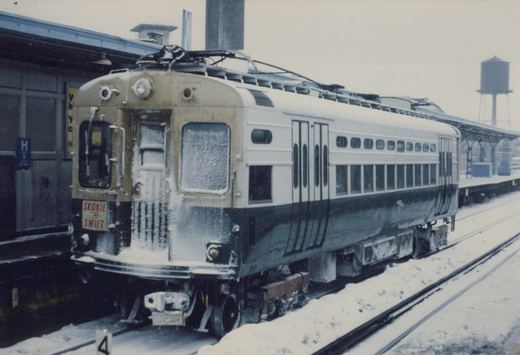 A snow-covered CTA 4000-series rapid transit car, number 23, labeled 'Skokie Swift,' is stopped at a station platform with a water tower visible in the background.
