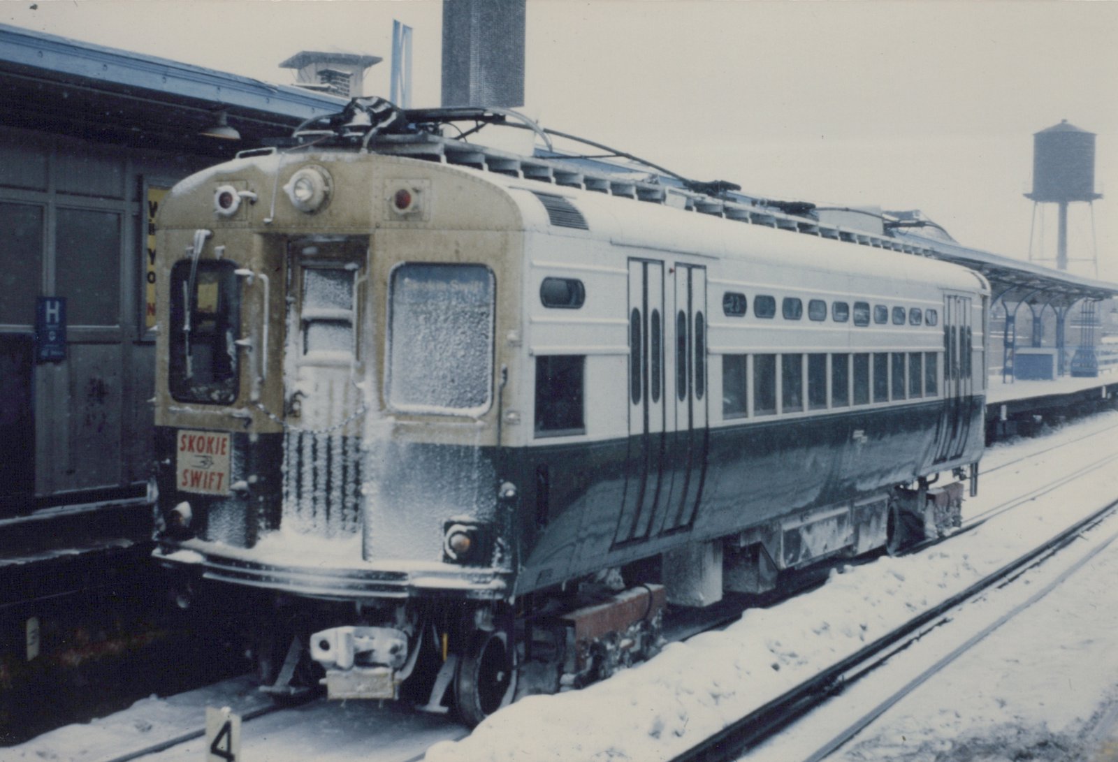 A snow-covered CTA 4000-series rapid transit car, number 23, labeled 'Skokie Swift,' is stopped at a station platform with a water tower visible in the background.