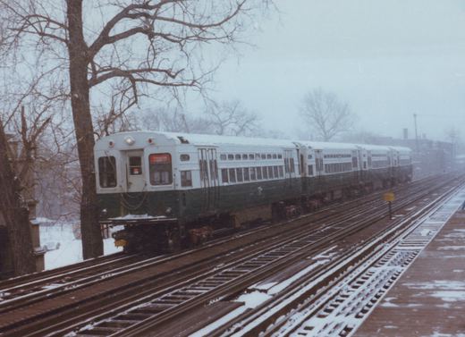 A green and white electric train labeled 'EVANSTON EXPRESS' moves along snow-dusted tracks beside bare trees on a cloudy winter day.