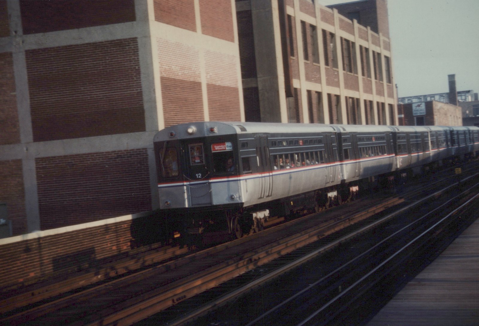 A silver CTA train with red and blue stripes, numbered 12, travels on an elevated track beside a large brick building on a clear day.