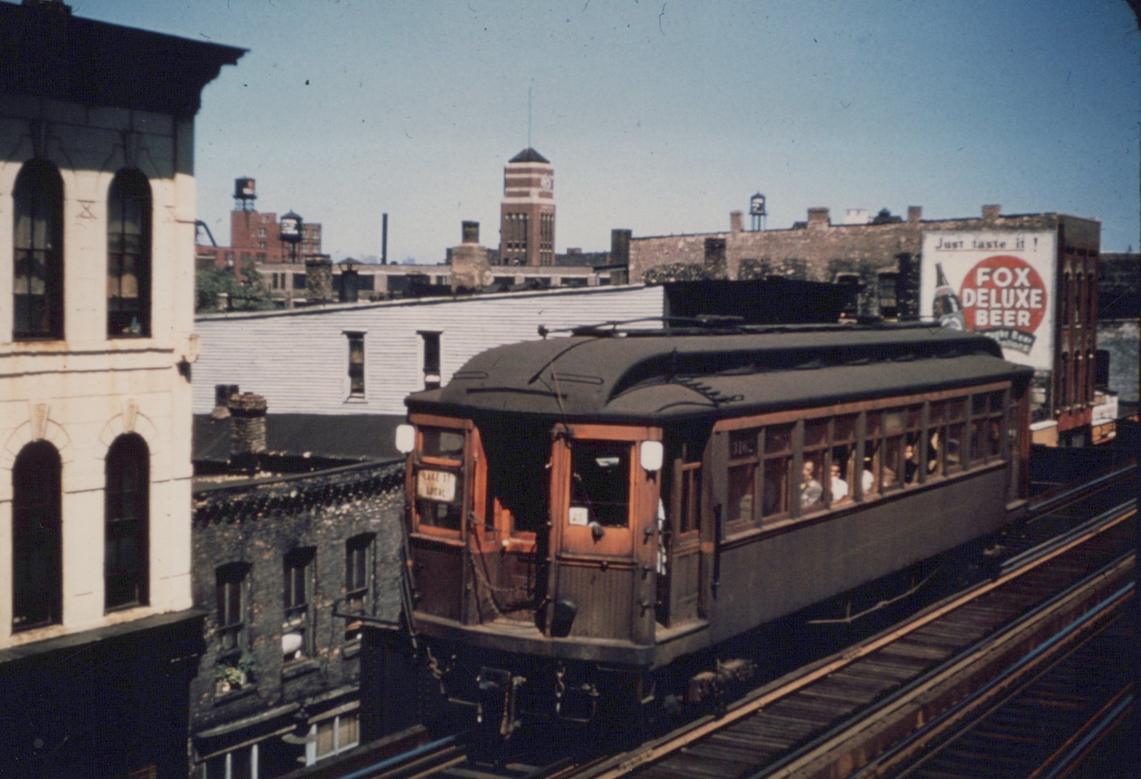 A vintage interurban streetcar with the number 25, in dark green and wood trim, runs on an elevated track through a city neighborhood with a 'Fox Deluxe Beer' billboard in the background.