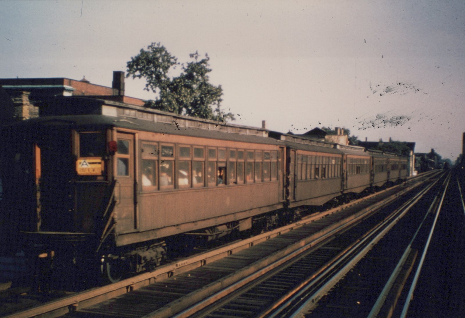 An old, light-colored wooden interurban car with many windows travels on elevated tracks alongside buildings and trees under an overcast sky.