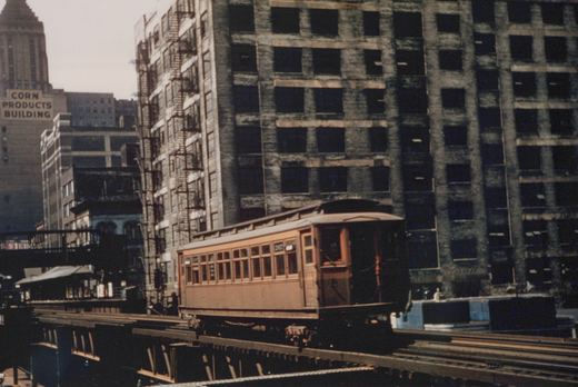A vintage wooden rapid transit car on an elevated track in a city, with tall buildings in the background.