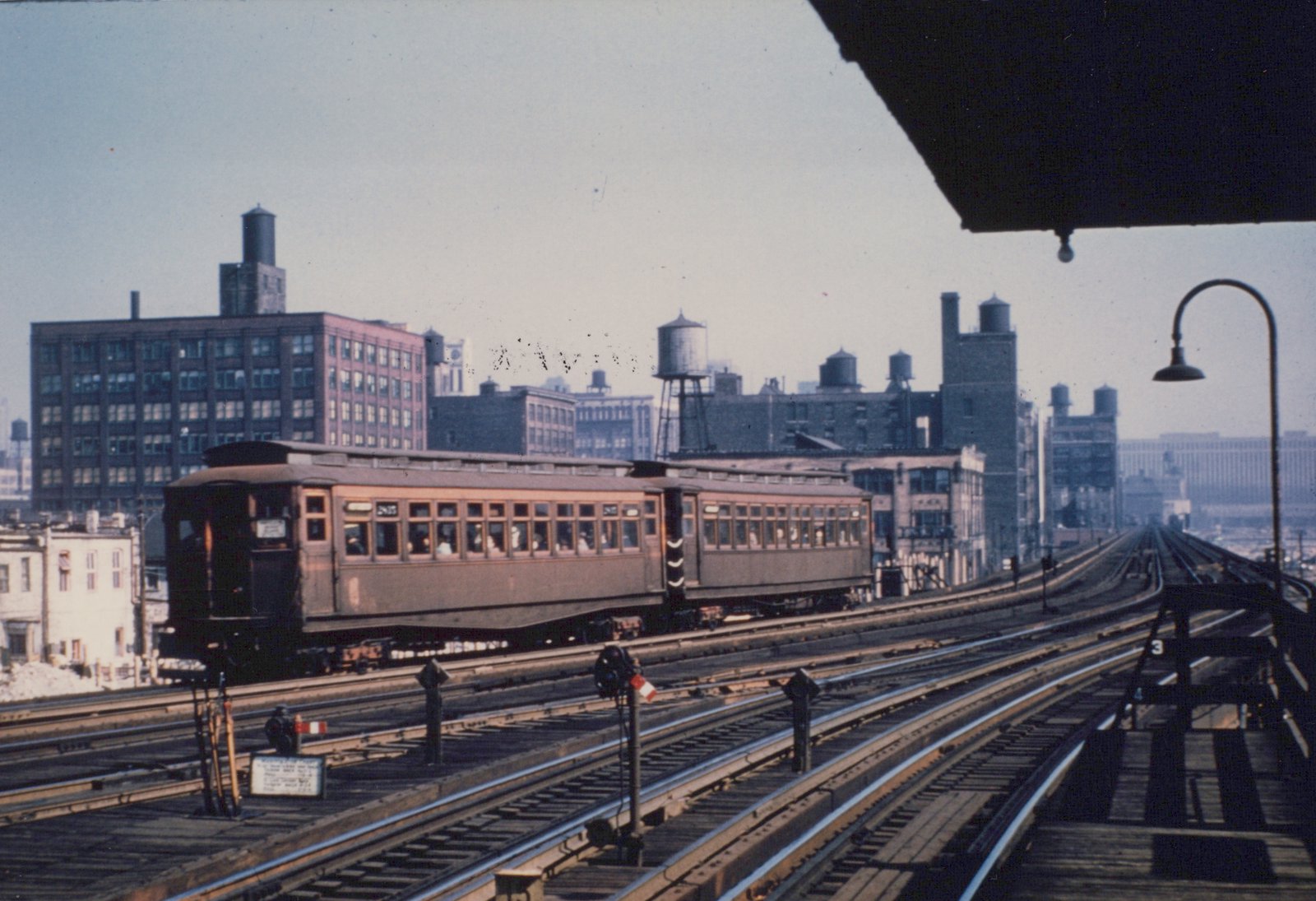 A vintage interurban car running on an elevated railway track, with city buildings and water towers in the background.