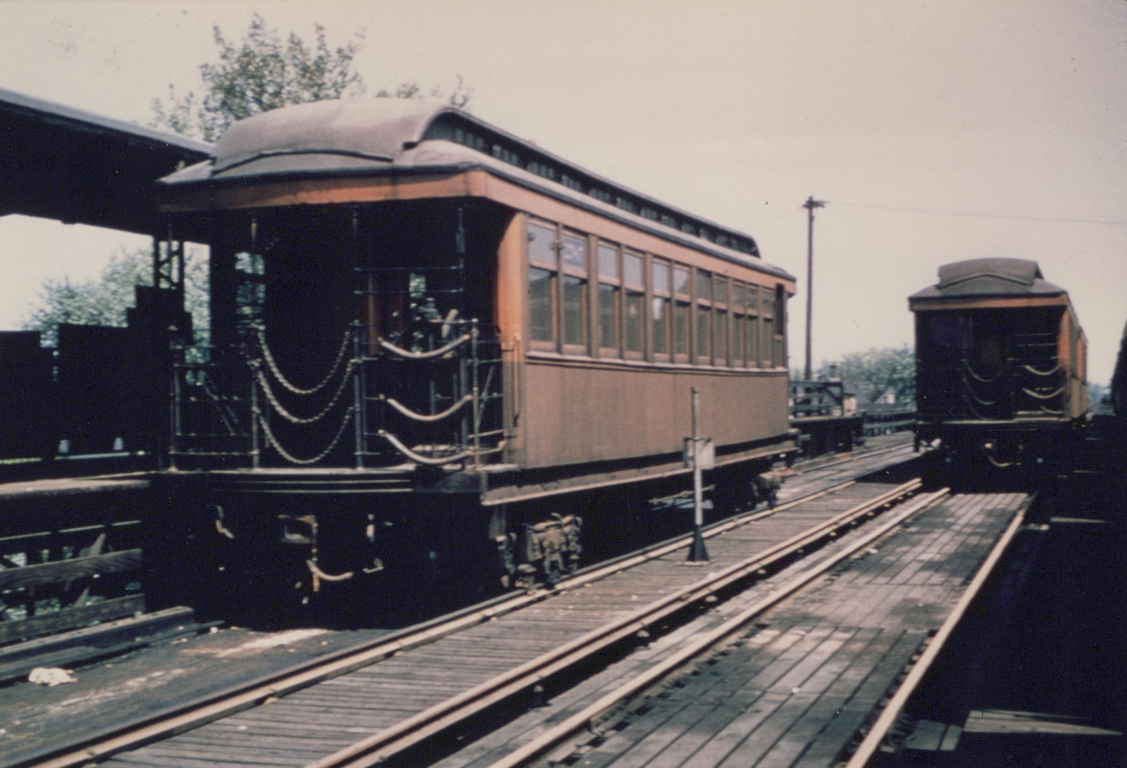 Two vintage wooden interurban cars in brown and cream livery are parked on an elevated wooden platform, with overhead wires visible.