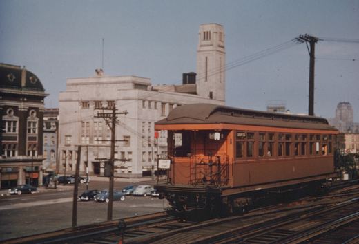 An orange and brown interurban railcar on an elevated track, with a sign reading 'LOGAN SQ' and 'LOGAN SQUARE', set against a backdrop of 1940s Chicago buildings and parked cars.