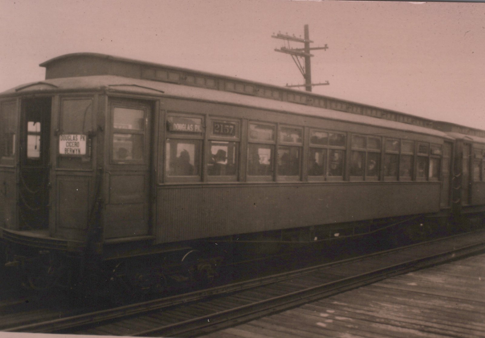 A black-and-white photo of a vintage Chicago Aurora and Elgin interurban car, number 2157, on an elevated track with passengers visible inside; signs indicate routes to Douglas Park, Cicero, and Berwyn.