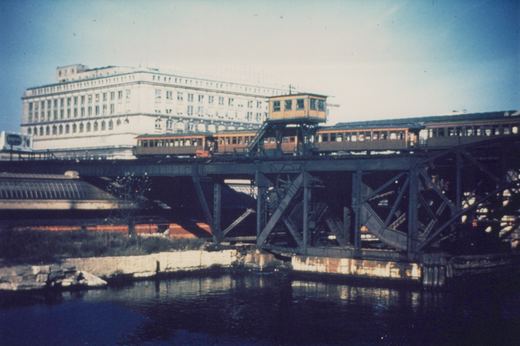An elevated train of vintage wooden 'L' cars travels across a metal truss bridge above a river, with a large building visible behind it under a clear sky.