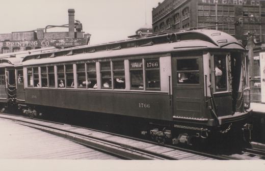Black and white photo of a vintage Chicago interurban car, number 1766, labeled 'Evanston Express,' stopped on city street tracks with passengers inside and commercial buildings behind.