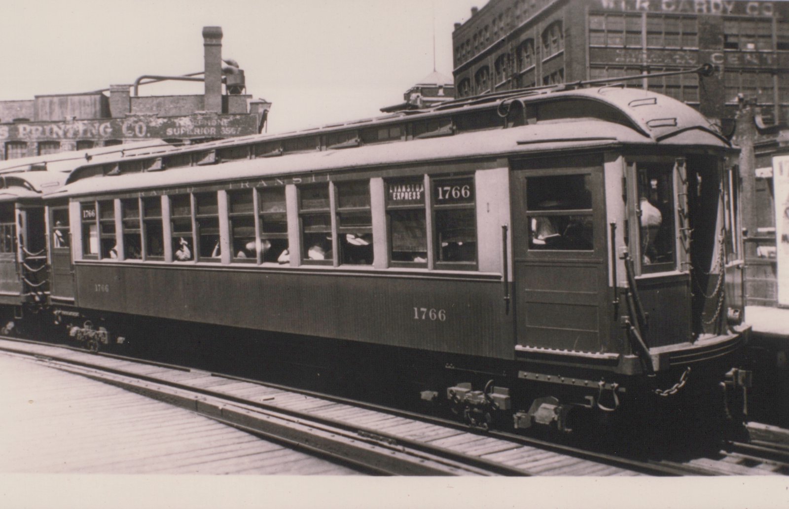 Black and white photo of a vintage Chicago interurban car, number 1766, labeled 'Evanston Express,' stopped on city street tracks with passengers inside and commercial buildings behind.