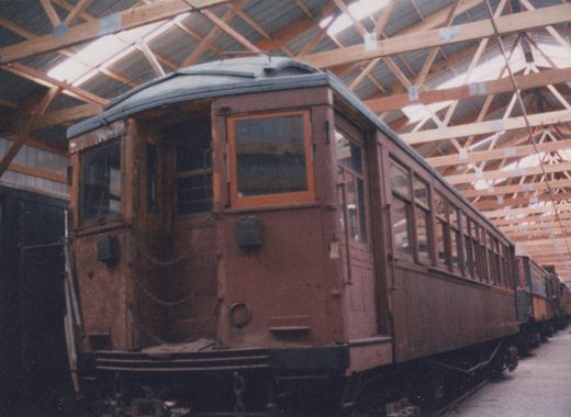 A vintage wooden interurban train car is parked inside a large wooden building with a truss roof; other similar cars are visible behind it.