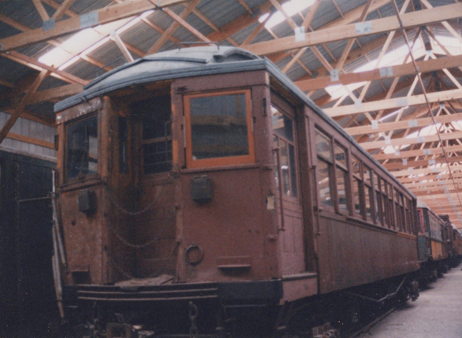 A vintage wooden interurban train car is parked inside a large wooden building with a truss roof; other similar cars are visible behind it.