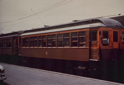 Side view of two orange and maroon interurban cars on a street, with overhead wires and a vintage car in the corner.