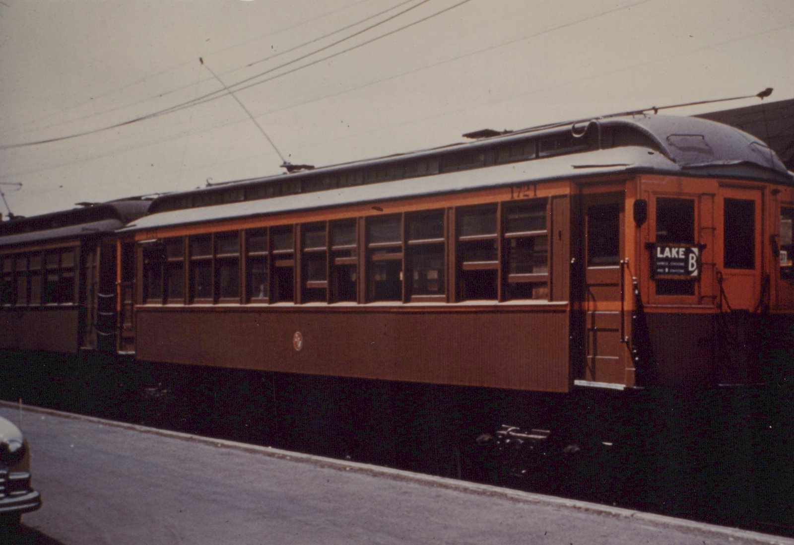 Side view of two orange and maroon interurban cars on a street, with overhead wires and a vintage car in the corner.
