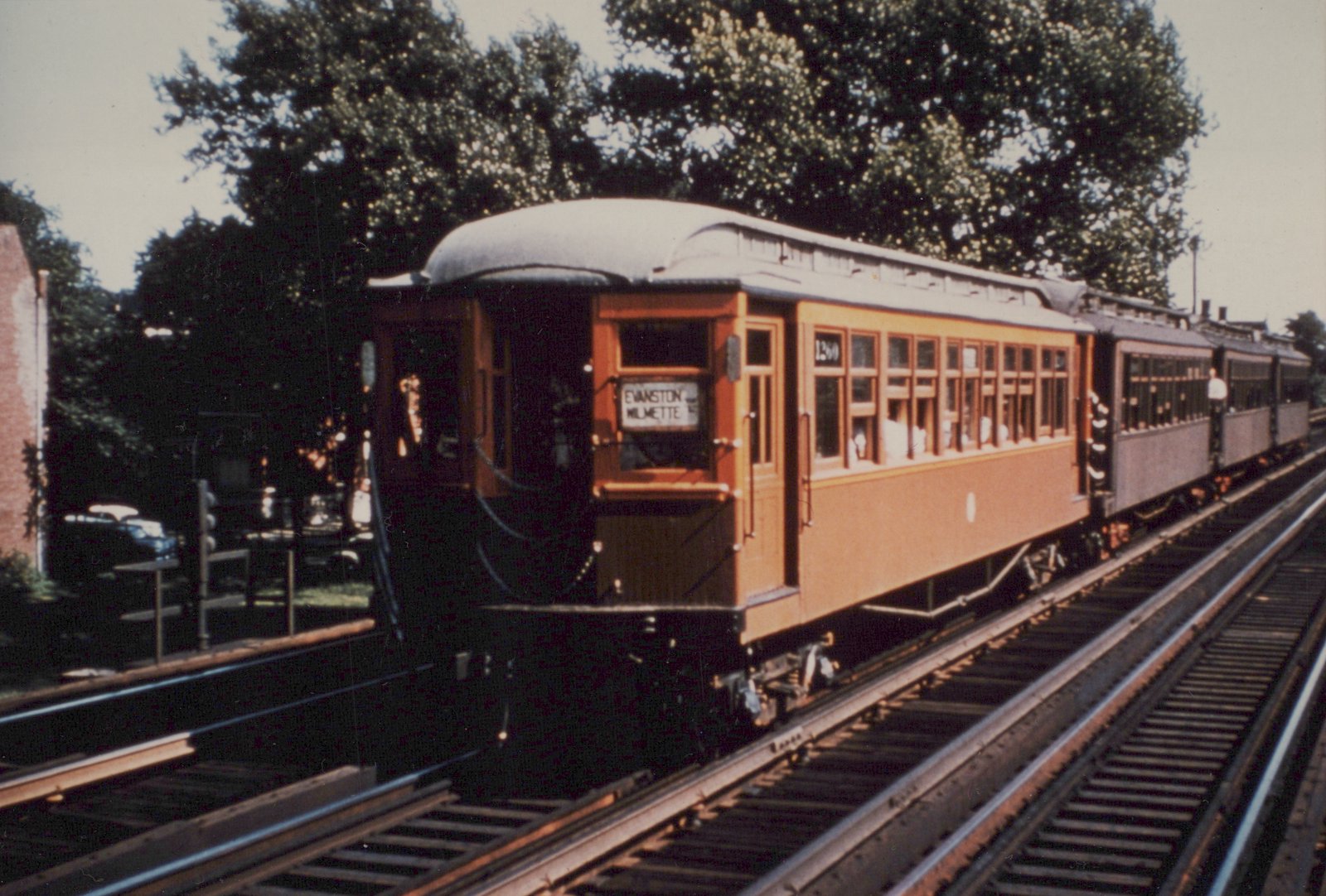 A vintage orange interurban train with the number 1260 on its side and a destination sign reading 'EVANSTON MILMETTE' travels on elevated tracks surrounded by trees on a clear day.