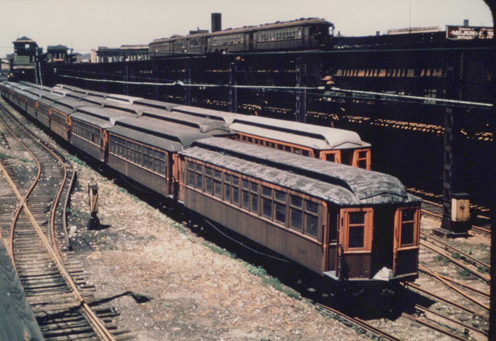 A vintage wooden interurban train car numbered 1237 is parked on tracks in a rail yard, with a dark green and wood-trim livery, under clear skies.