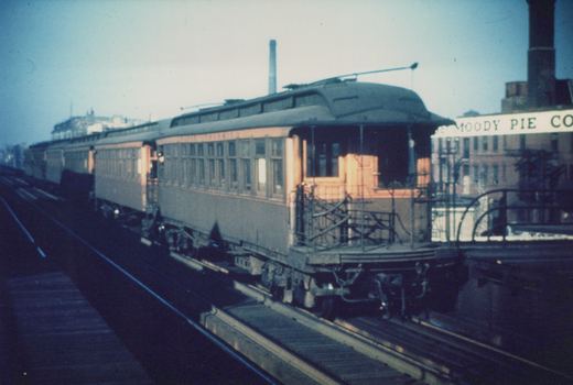 An old electric train car with 'RAPID TRANSIT' lettering on its side travels on elevated tracks near a building with a 'MOODY PIE CO' sign.