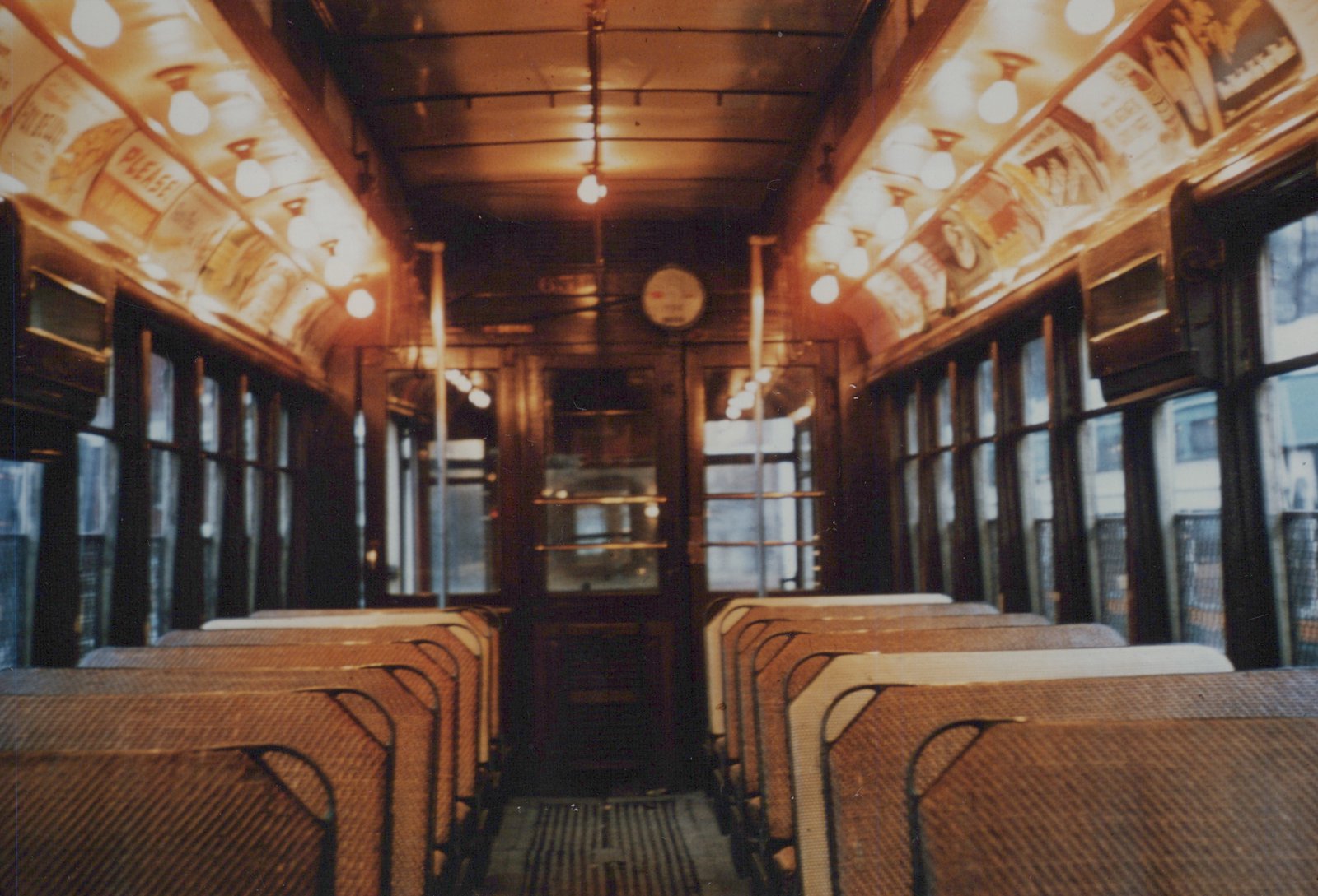 View from inside a vintage streetcar, looking down the aisle towards the front door, with rows of woven seats and wooden paneling on the walls and ceiling.