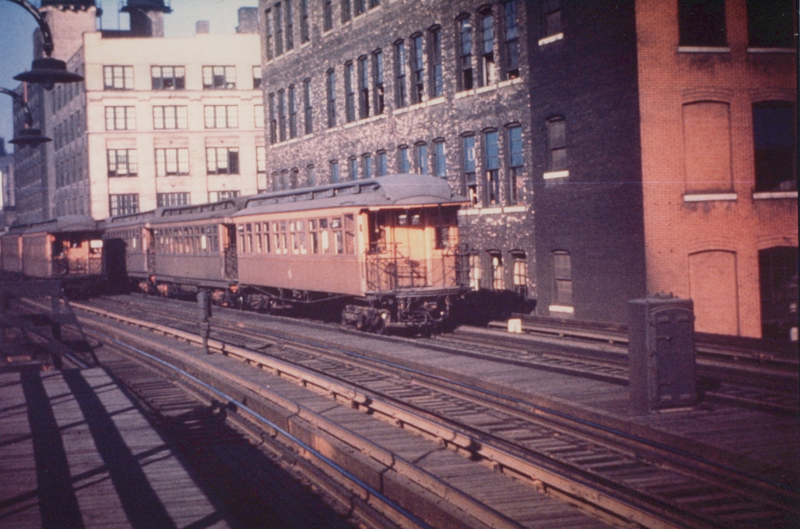 A three-quarter front view of a train of old, wood-bodied elevated cars on an elevated track in Chicago, with large brick and stone industrial buildings in the background under a clear sky.