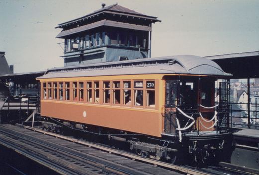 A vintage orange interurban car numbered 270 is parked on an elevated railway platform with a control tower in the background.