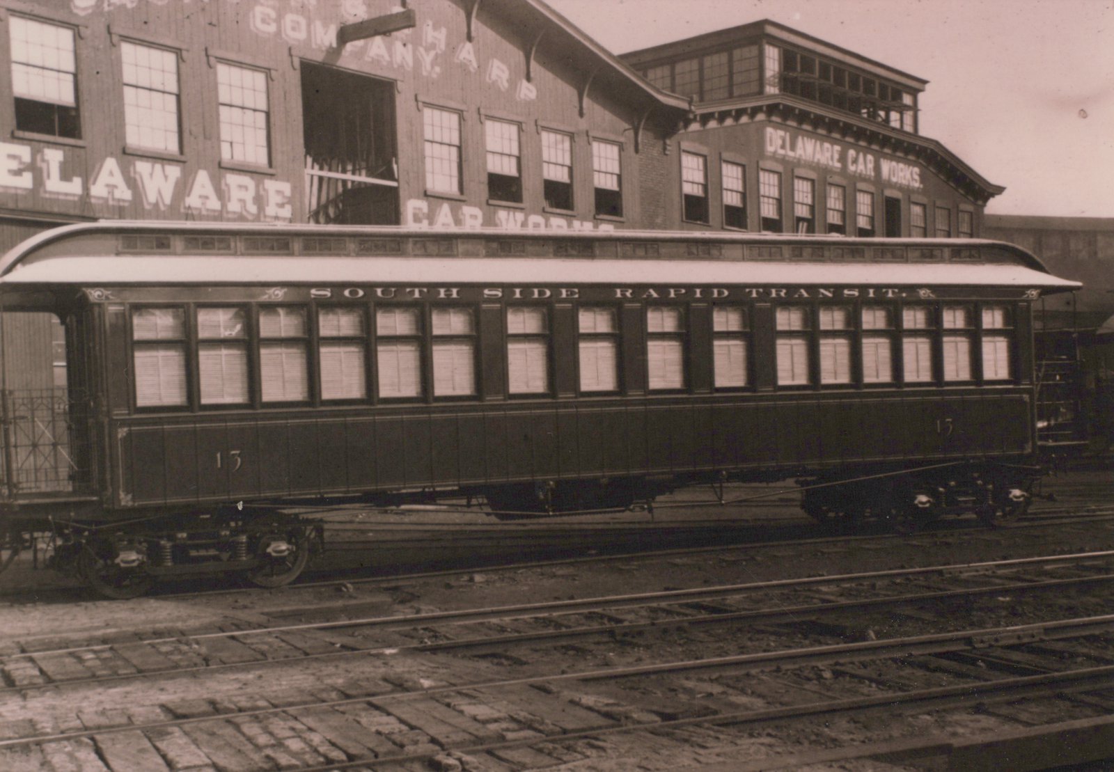A side view of a vintage rapid transit car labeled 'South Side Rapid Transit' with the number 13, parked in front of a building with 'Delaware Car Works' lettered on it in 1892.