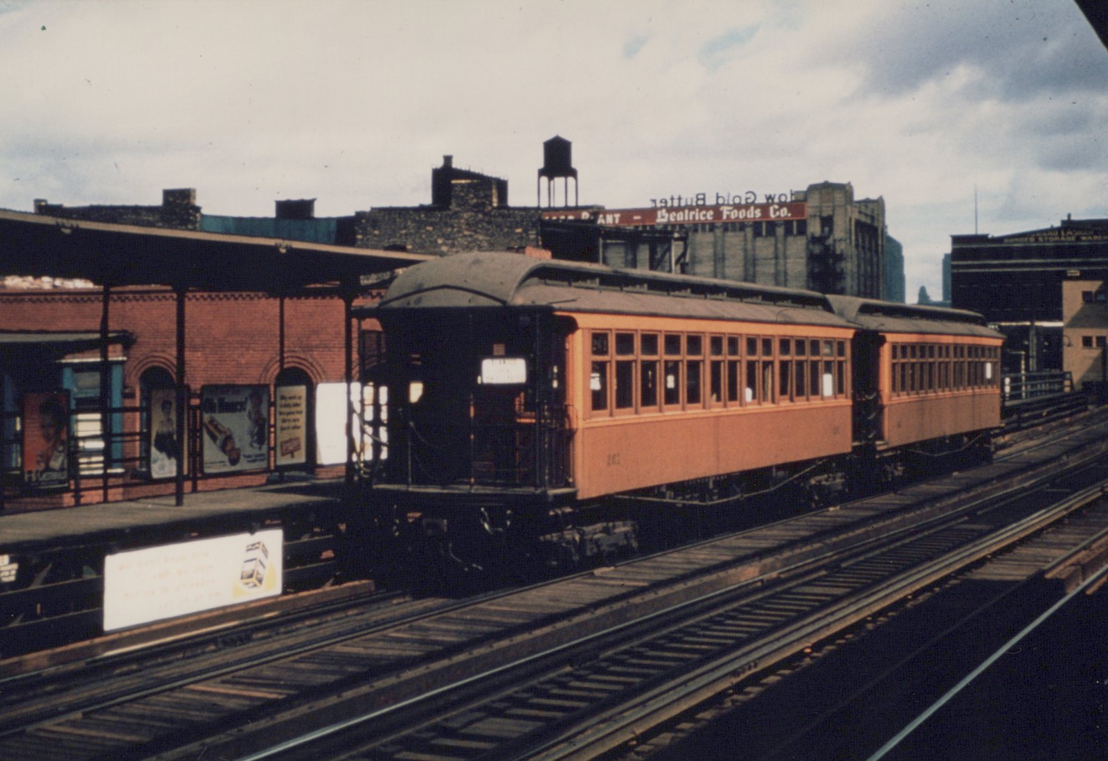 An orange and black interurban car with the number 261 is stopped at a brick elevated station platform, with a large 'Beatrice Foods Co.' billboard visible on a building in the background.