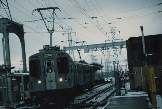 A two-car train of Chicago Transit Authority 2600-series rapid transit cars, with car 3445 in the lead, is stopped at an elevated station platform in winter, with snow on the ground and tracks.