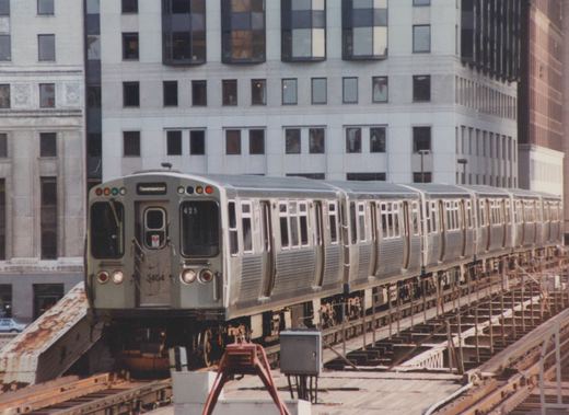 A silver and gray Chicago Transit Authority train, number 3404, travels along an elevated track over a city street, with a modern building in the background.
