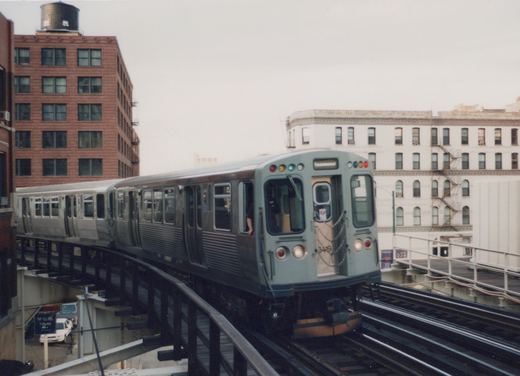 A silver Chicago Transit Authority rapid transit train, numbered 3419, travels along an elevated track in a city, with buildings in the background under an overcast sky.