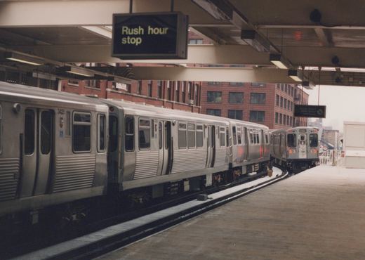 Side view of a stainless steel Chicago Transit Authority train at an elevated station platform in winter, with another train visible on an adjacent track and a sign above reading 'Rush hour stop'.