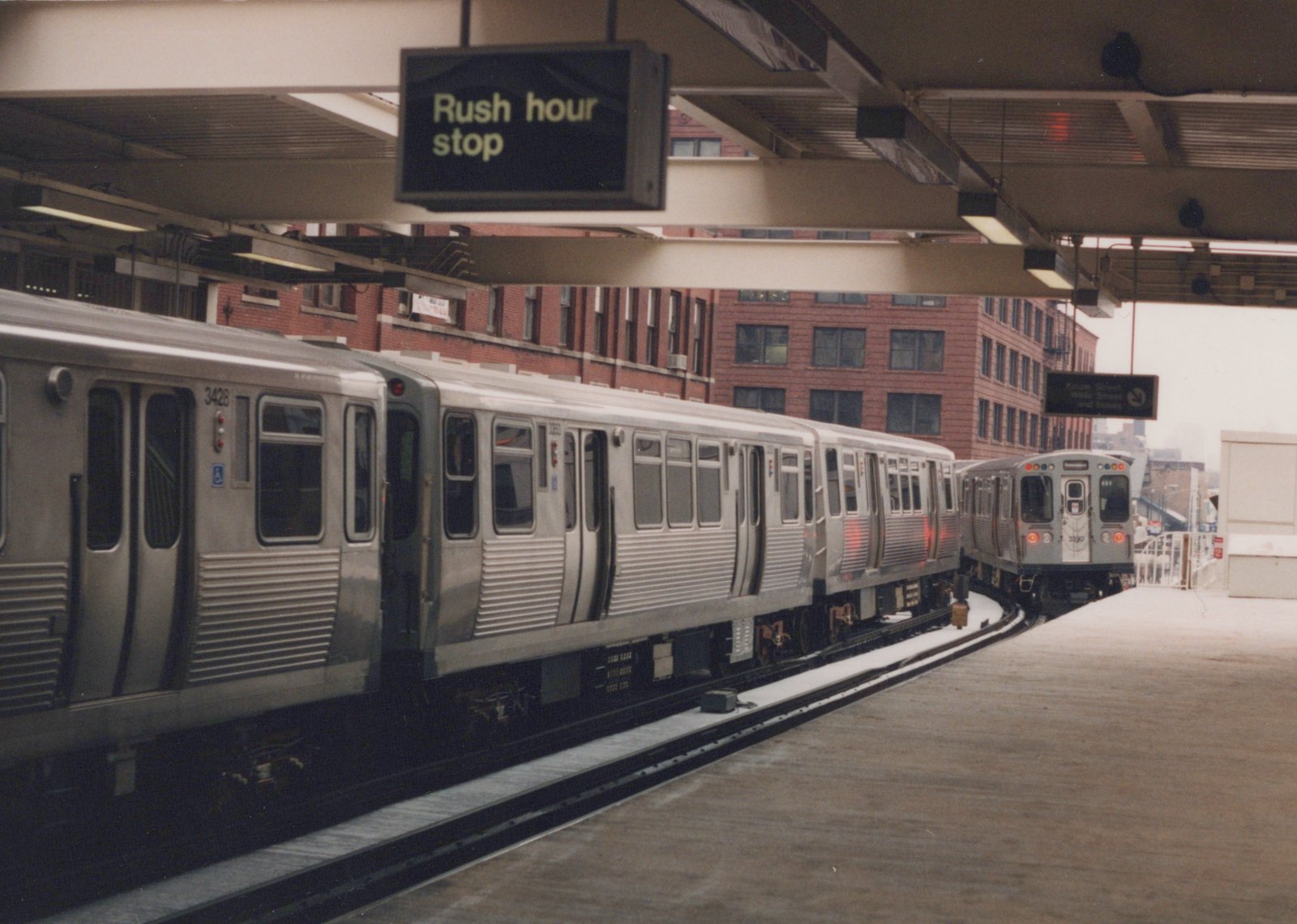 Side view of a stainless steel Chicago Transit Authority train at an elevated station platform in winter, with another train visible on an adjacent track and a sign above reading 'Rush hour stop'.