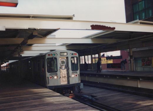 A light gray CTA train with the number 3366 on the front is stopped at an elevated station platform with a sign reading 'Merchandise Mart'. Three people are visible on the opposite platform.