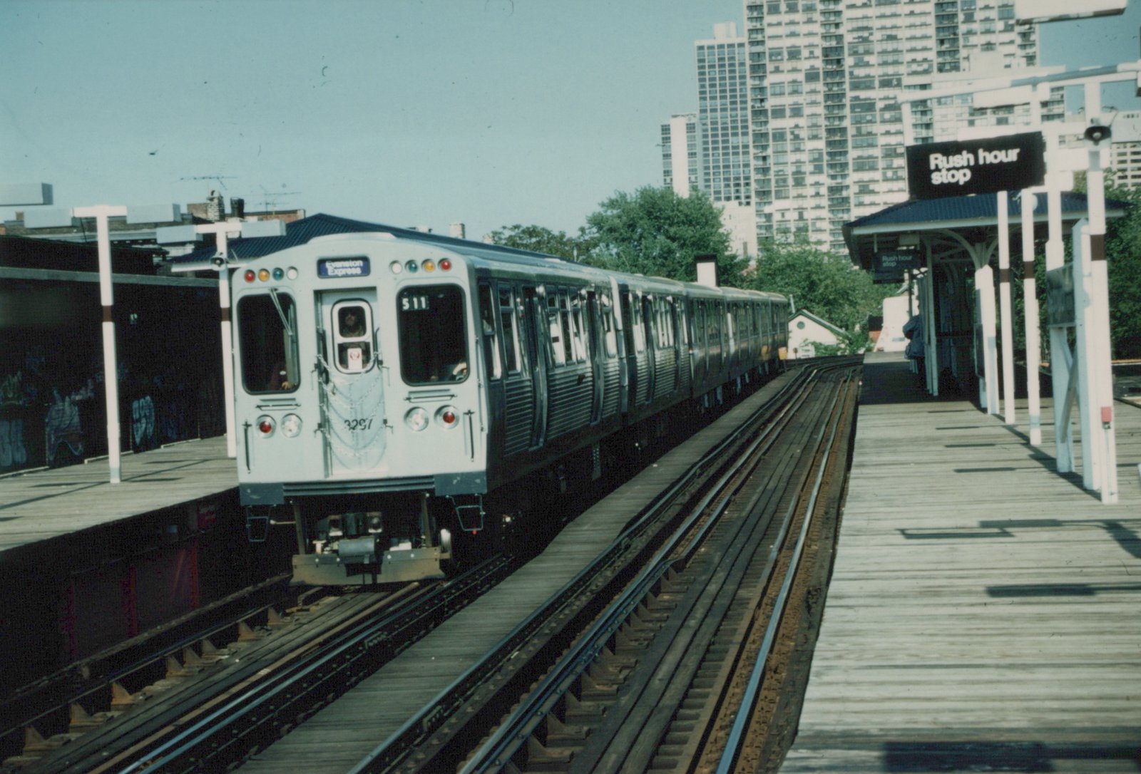 A white and blue CTA train is stopped at an outdoor elevated station with a sign that reads 'Rush hour stop'.