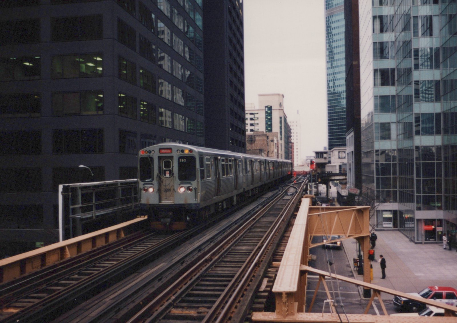 A silver Chicago Transit Authority train with the number 3289 and destination 'Midway' travels on elevated tracks between tall buildings on an overcast day.